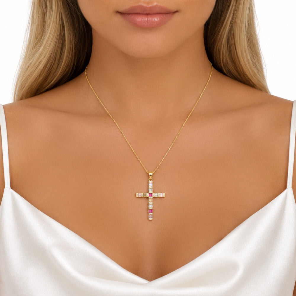A gold cross pendant necklace with clear and pink gemstones, worn by a model in a white satin top against a white background.