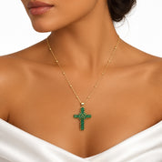 A gold cross pendant necklace with green gemstone accents, worn by a model in a close-up shot.