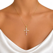 A gold cross pendant necklace with clear and pink gemstones, worn on a model's neck over a white satin top.
