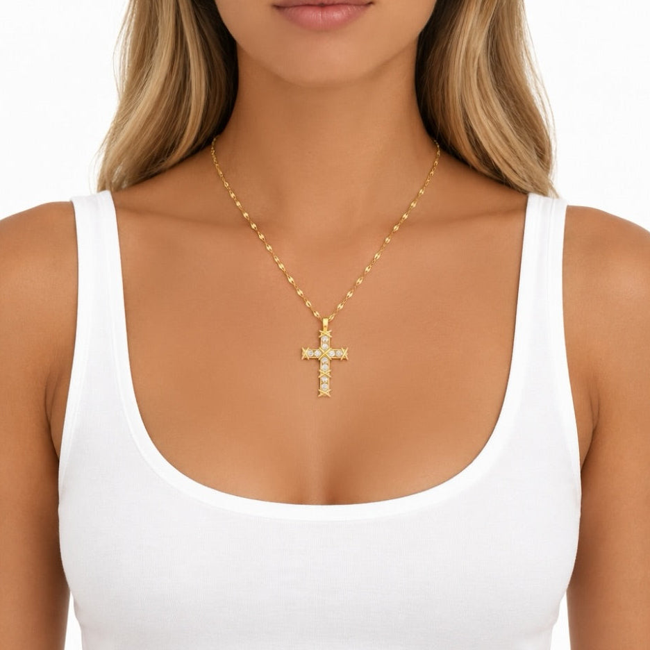 A gold cross pendant necklace with clear stones, worn by a model in a white tank top against a white background.