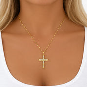 A gold cross pendant necklace with clear stones, worn by a model in a white top.
