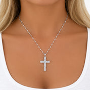 A silver cross pendant necklace with clear stones, worn on a model's neck over a white tank top.