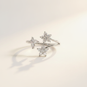 A delicate silver open ring featuring three flower-shaped clusters of clear stones, photographed on a clean, light beige background with soft shadows.