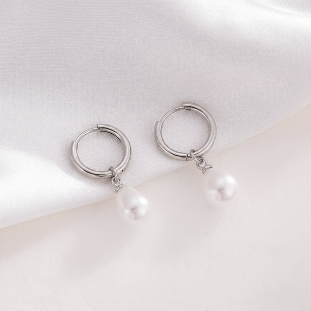 A pair of silver huggie hoop earrings with a small dangling white pearl, displayed on a soft white satin fabric background.