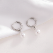 A pair of silver huggie hoop earrings with a small dangling white pearl, displayed on a soft white satin fabric background.