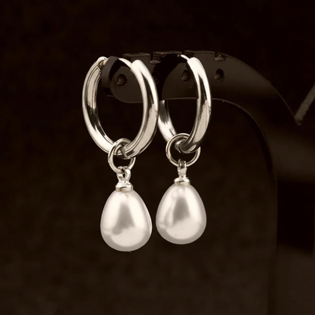 A pair of silver hoop earrings with dangling teardrop-shaped white pearls, displayed on a black jewelry stand against a dark brown background.