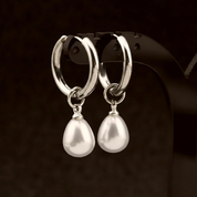 A pair of silver hoop earrings with dangling teardrop-shaped white pearls, displayed on a black jewelry stand against a dark brown background.