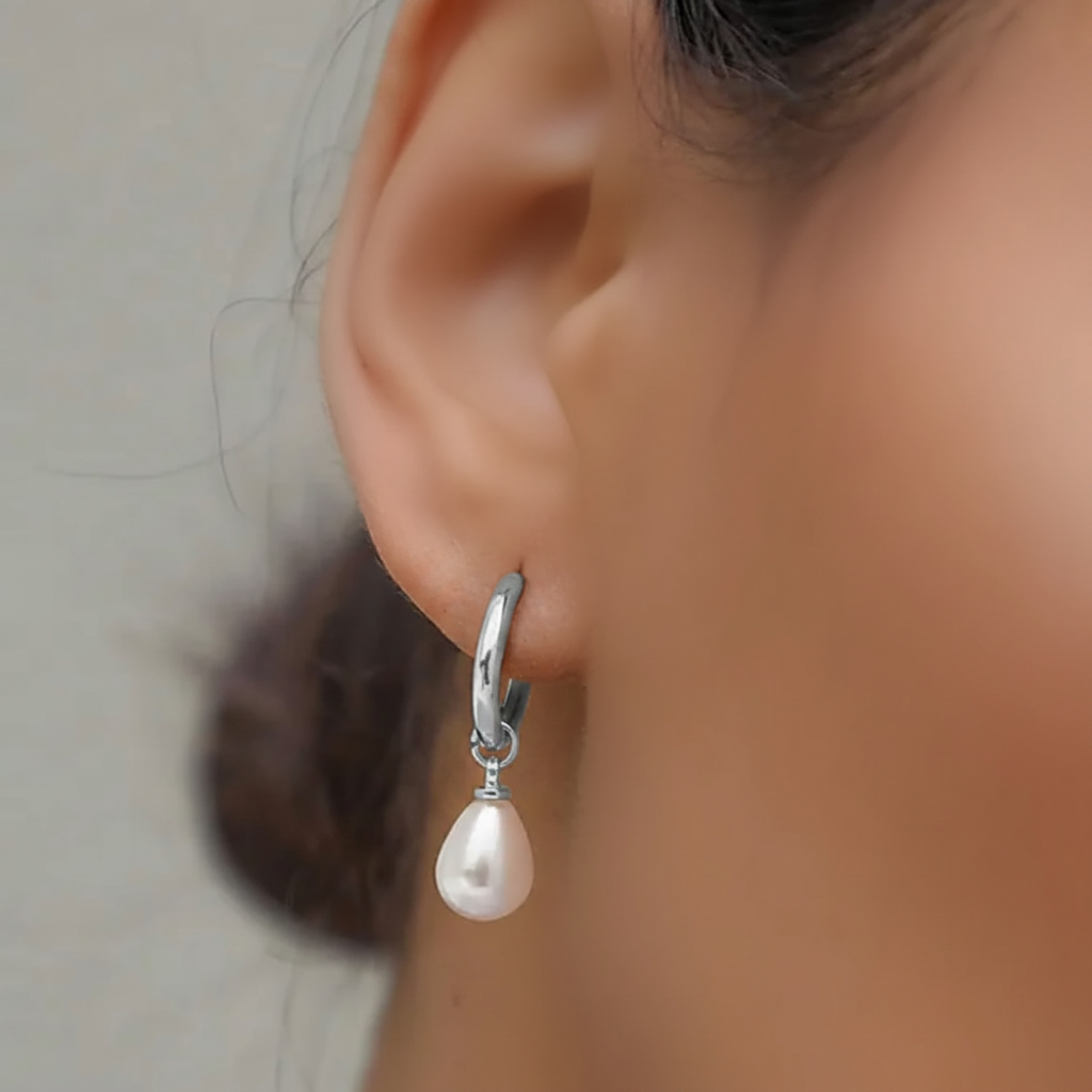 A close-up of a person's ear wearing a silver huggie hoop earring with a dangling teardrop-shaped white pearl.