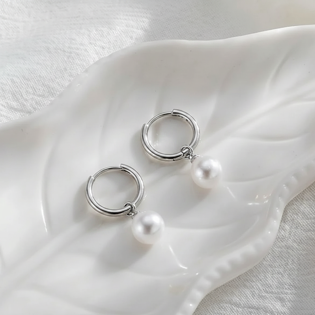 A pair of silver hoop earrings with dangling white pearl beads, displayed on a white ceramic leaf-shaped dish over a textured white fabric background.