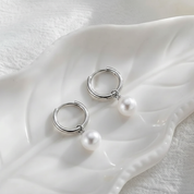A pair of silver hoop earrings with dangling white pearl beads, displayed on a white ceramic leaf-shaped dish over a textured white fabric background.